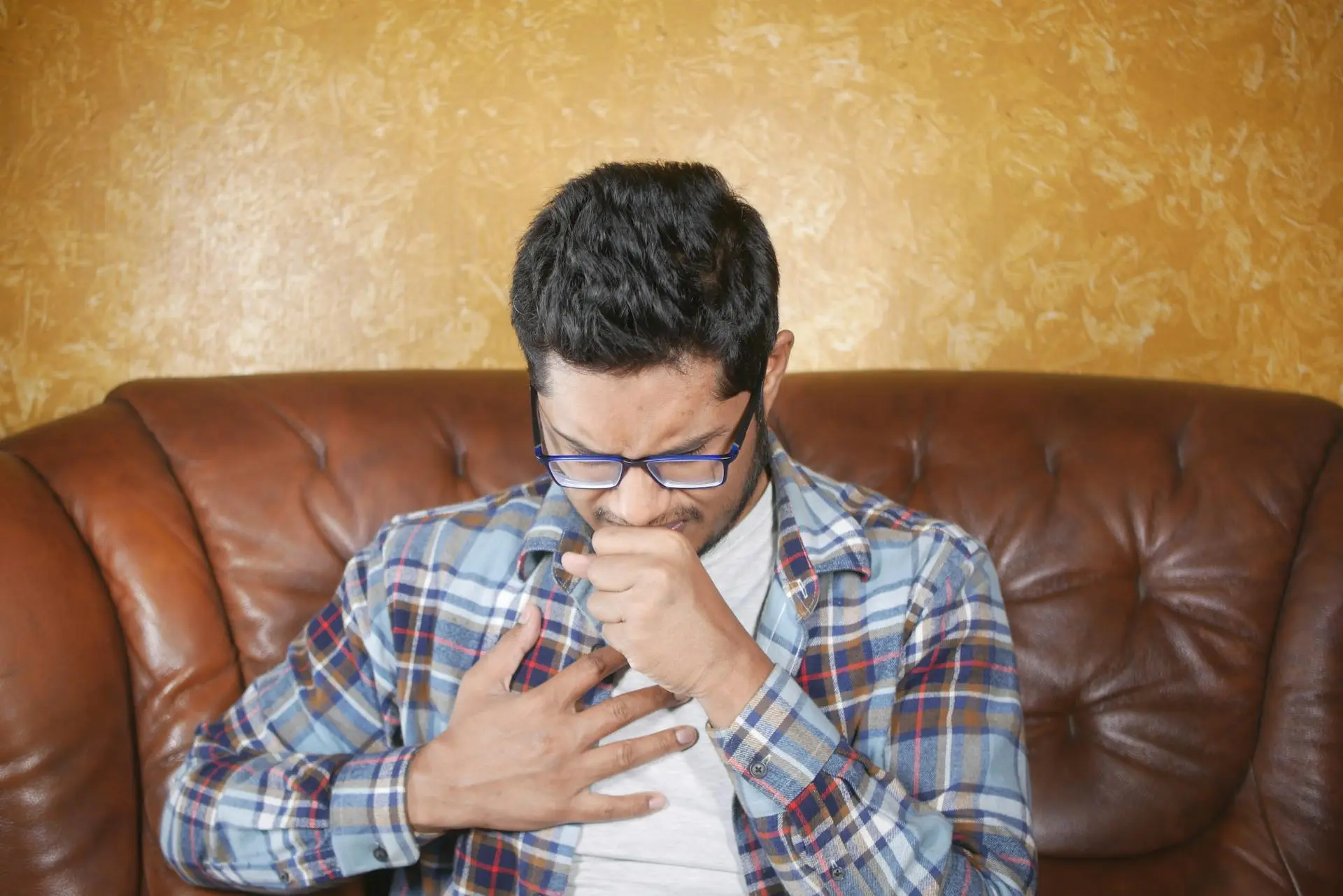 Man in plaid shirt coughing with hand to chest on brown couch